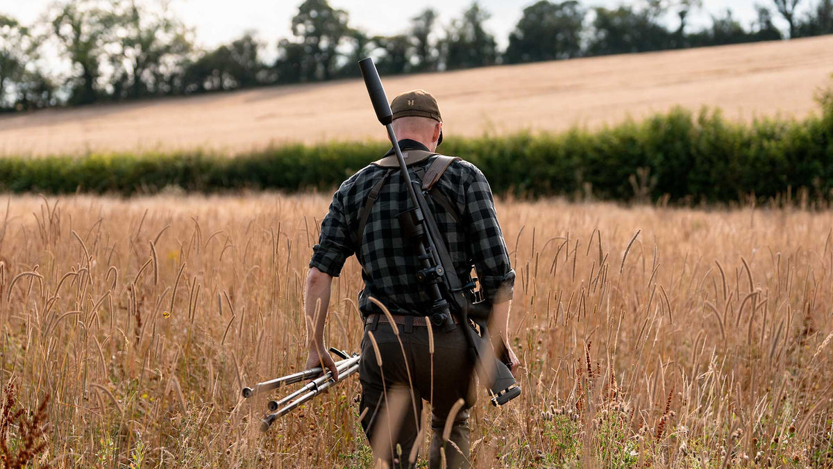 Chasseur de dos marchant dans les hautes herbes, carabine équipée de la Pulsar Thermion 3 à l’épaule et canne de tir à la main