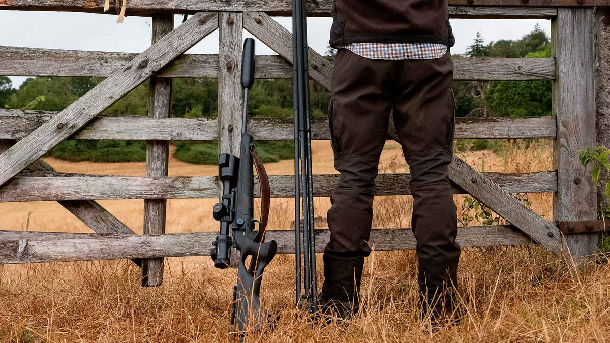 Carabine équipée de la lunette thermique Pulsar Thermion 3 posée contre une barrière en bois, à côté d’un chasseur et de sa canne de tir
